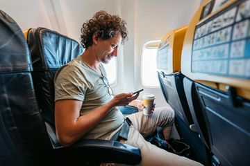Handsome young man using smart phone and drinking coffee while in an Airplane.