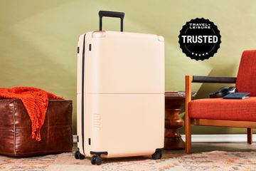 July Checked Plus suitcase displayed between a leather floor cushion and a chair