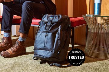A Kuhl backpack sits on the ground next to a person's legs