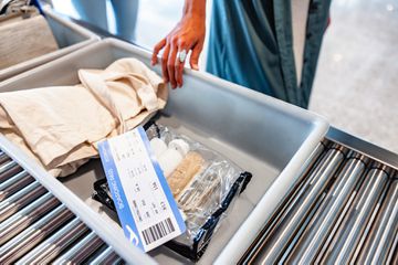 Grey tray filled with liquid toiletries in a clear zip top bag, boarding pass and a beige tote bag. A female hand is holding the tray on a roller conveyors table at airport security.